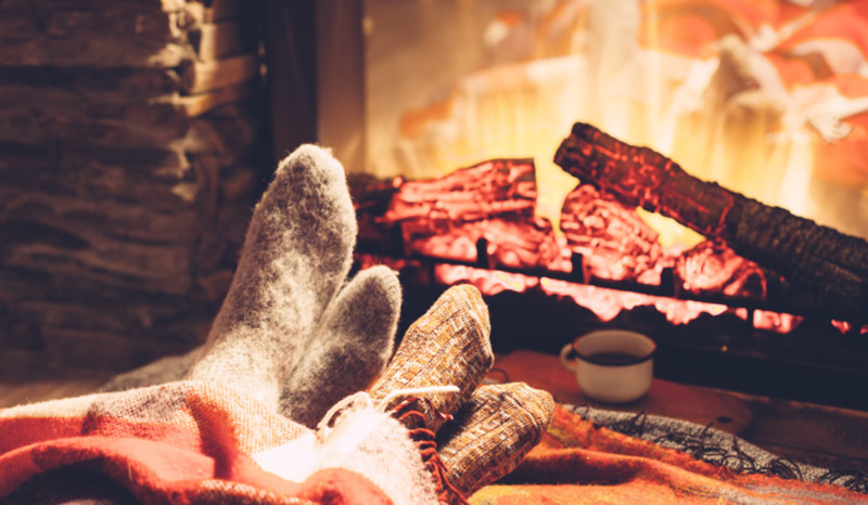 Die besten Tipps zum Gas sparen Die Füße zweier Personen sind eingekuschelt in dicken Socken und mit einer Wolldecke vor dem Kamin.