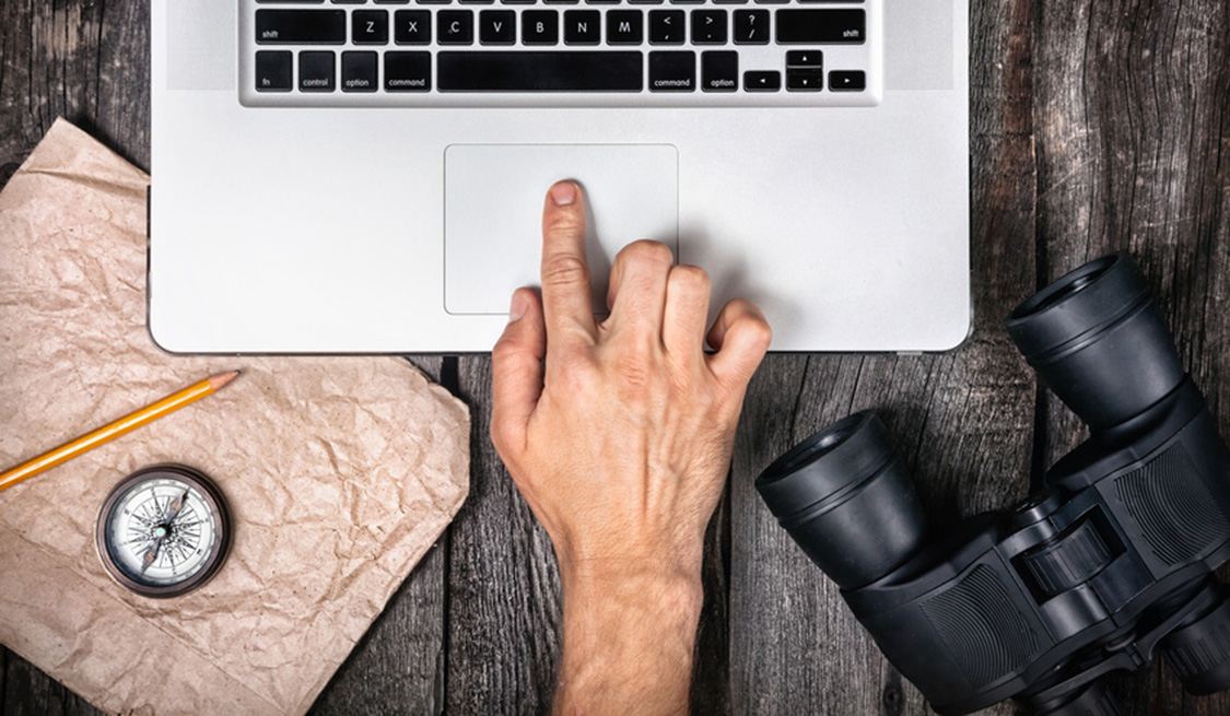 Mann hält einen Finger auf das Mousepad seines Laptops. Daneben liegen Kompass und Fernglas.