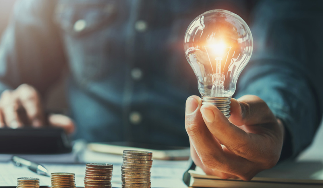 Der Kilowattstundenpreis | Verbraucherthemen Mann hält leuchtende Glühlampe in der Hand. Daneben ein paar Stapel Geldmünzen