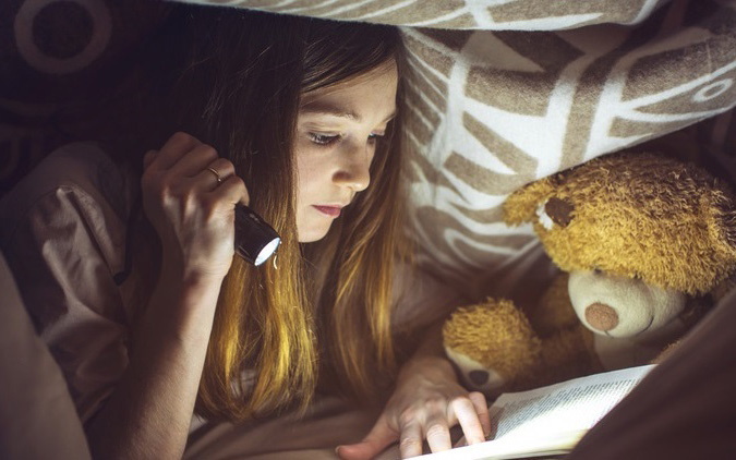 Mädchen Liegt mit Taschenlampe und Buch unter der Bettdecke.