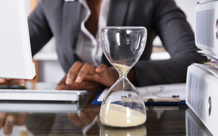 Frau im Büro am Schreibtisch sitzend. Neben Aktenordnern steht eine Sanduhr aus Glas