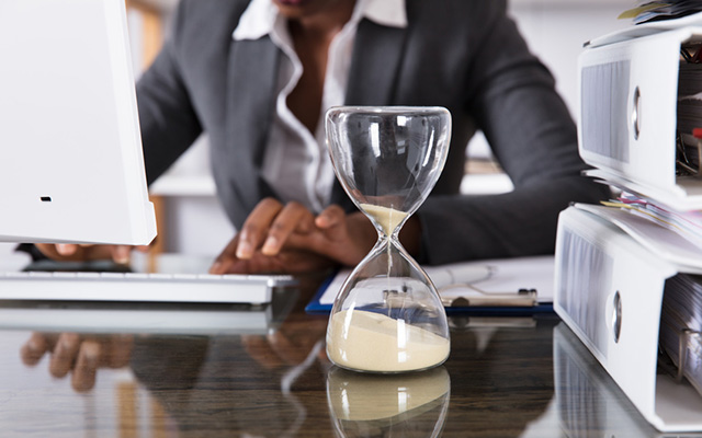 Frau im Büro am Schreibtisch sitzend. Neben Aktenordnern steht eine Sanduhr aus Glas