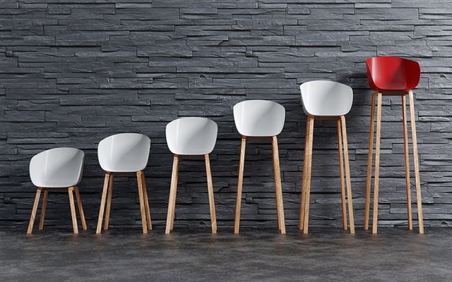 Fünf weiße und ein roter Stuhl stehen nebeneinander mit immer länger werdenden Stuhlbeinen vor einer grauen Mauer
