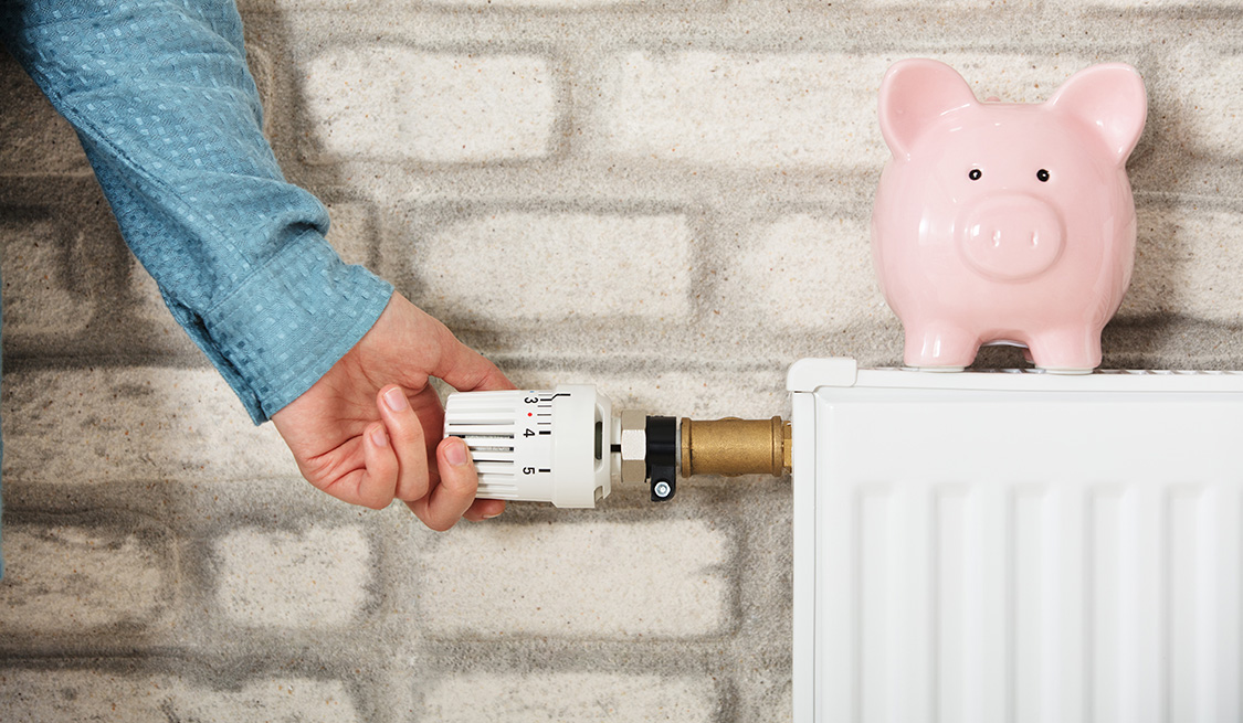 Zentralheizung | Strom und Gas | Lexikon Eine Person hat die Hand am Temperaturregler einer Gasheizung. Auf der Heizung steht ein rosa Sparschwein.