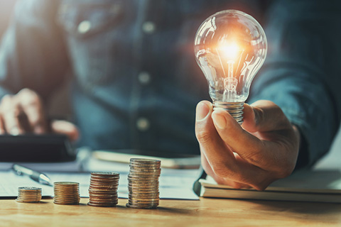 Über Uns | cheapenergy24 Mann hält leuchtende Glühlampe in der Hand. Daneben ein paar Stapel Geldmünzen
