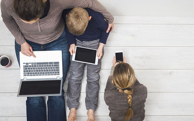 Frau sitzt mit zwei Kindern auf dem Fußboden. Sie nutzen Laptop, Tablet und Smartphone