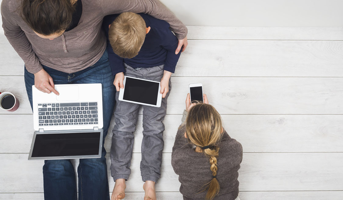 Was tun bei hohem Stromverbrauch? – Verbraucherfragen Frau sitzt mit zwei Kindern auf dem Fußboden. Sie nutzen Laptop, Tablet und Smartphone
